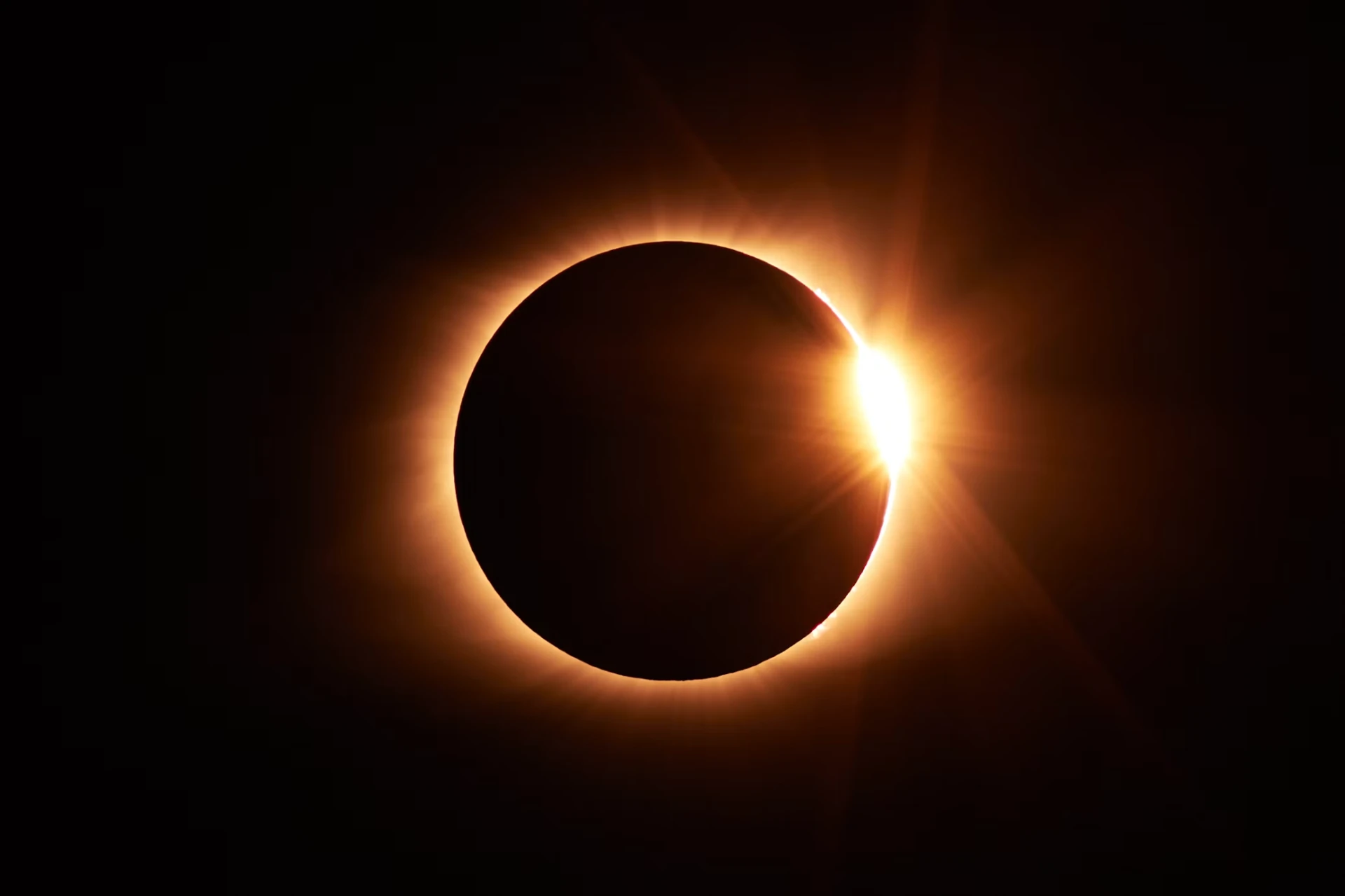 A solar eclipse with the Sun's corona glowing around the Moon and sunlight breaking through at the edge