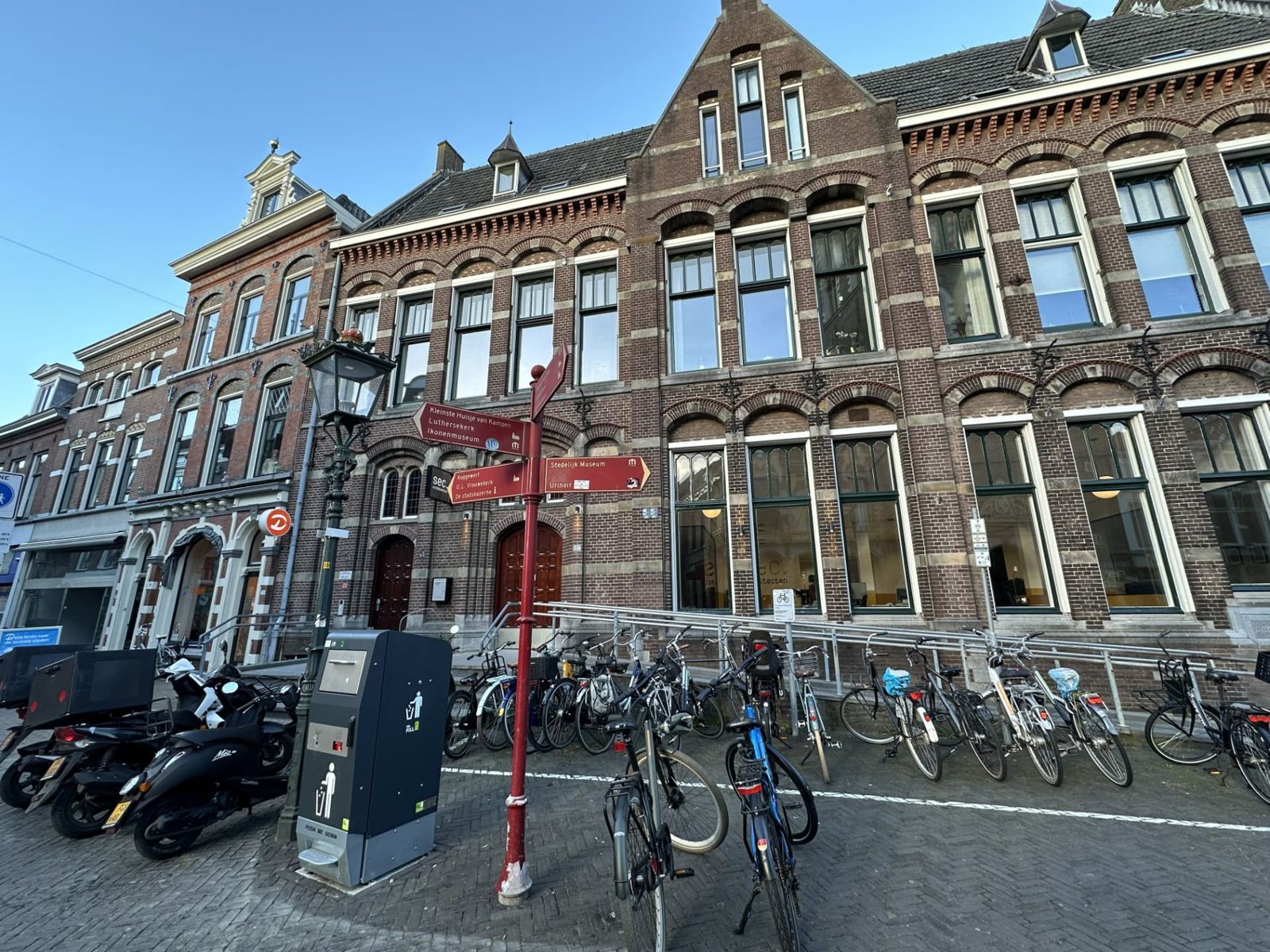 Dutch street with a red signpost pointing to local museums and churches, brick townhouses, and parked bicycles and scooters