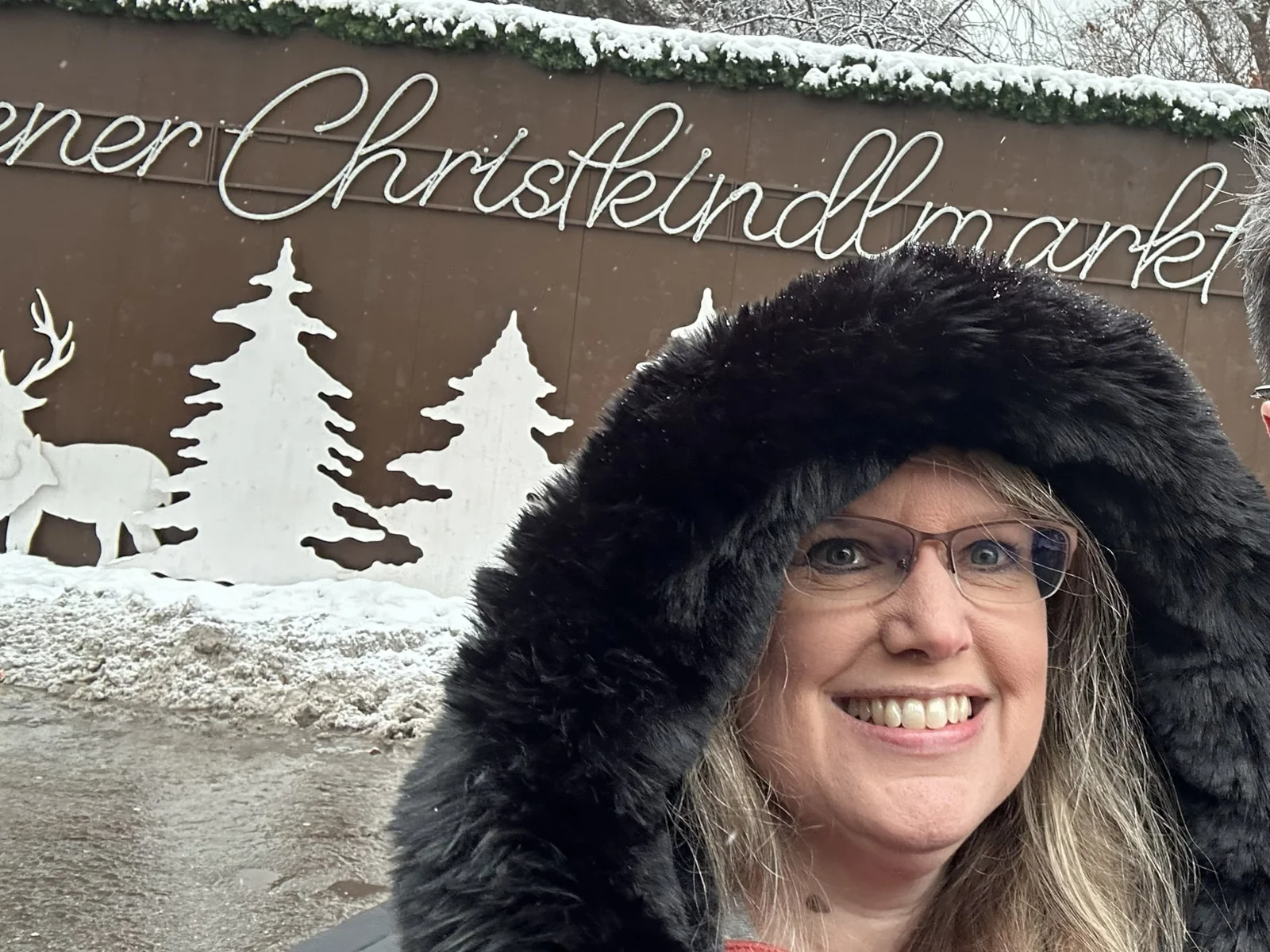 Melissa Yetter in a fur trimmed hood smiling in front of a snowy Christkindlmarkt sign.