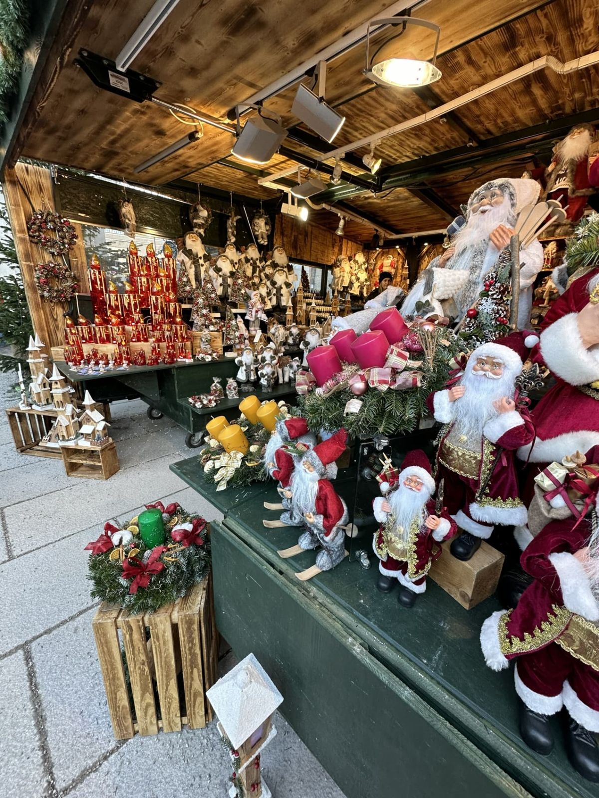Wooden Christmas market stall filled with Santa figures, red, pink, and yellow candles, evergreen wreaths, and small wooden village houses