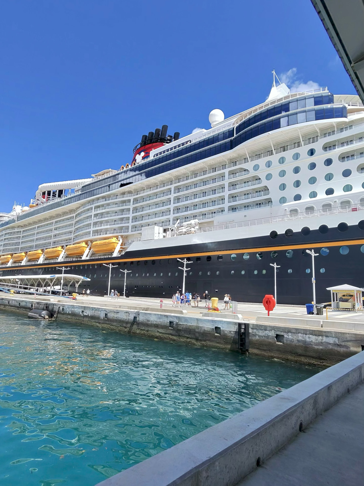 Disney cruise ship docked beside clear blue water under a cloudless sky.